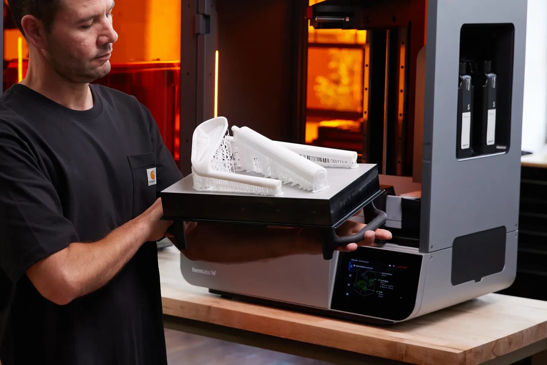 Operator holding a build platform with a white printed part beside an open Form 4L printer.